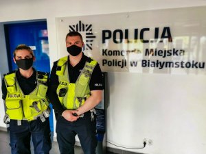 Policjantka i policjant z założonymi maseczkami. Mundurowi stoją na tle ściany z napisem Komenda Miejska Policji w Białymstoku