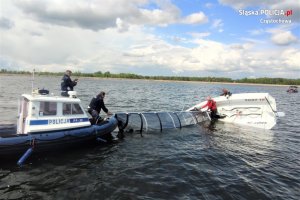 Policyjna łódka na której stoi dwóch policjantów, jeden z nich trzyma żaglówkę