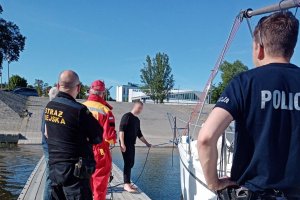 przystań dla łodzi, na pomoście umundurowany policjant, strażnik miejski, ratownik WOPR oraz mężczyzna trzymający linę dołączoną do łodzi, łódź stoi przy pomoście&quot;&gt;