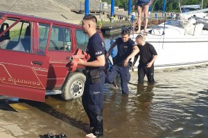 przystań dla łodzi, na pochylni prowadzącej do nurtu stoi samochód, do którego przyczepiona jest żaglówka, dwójka policjantów oraz mężczyzna ubrany na czarno brodzą w płytkiej wodzie&quot;&gt;