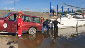 przystań dla łodzi, na pochylni prowadzącej do nurtu stoi samochód, do którego przyczepiona jest żaglówka, dwójka policjantów oraz mężczyzna ubrany na czarno brodzą w płytkiej wodzie, na lądzie stoi ratownik WOPR&quot;&gt;