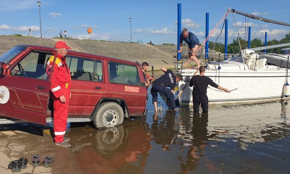 przystań dla łodzi, na pochylni prowadzącej do nurtu stoi samochód, do którego przyczepiona jest żaglówka, dwójka policjantów oraz mężczyzna ubrany na czarno brodzą w płytkiej wodzie, na lądzie stoi ratownik WOPR&quot;&gt;