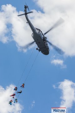 Policyjny Black Hawk w locie, pod nim na dwóch długich linach zwisa 6 ratowników GOPR.