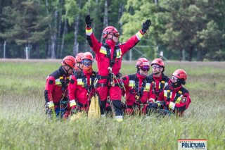 Grupa strażaków na łące, jeden daje znaki pilotom, pozostali klęczą.