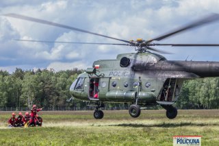 Policyjny Mi-8 ląduje na trawiastym lądowisku, oczekują na niego strażacy.