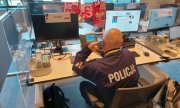 policjant przy biurku rozmawia przez telefon, w tle widać monitory komputerów, szalik z barwami narodowymi