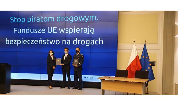 Na scenie stoją trzy osoby, które prezentują dokumenty. Od lewej Joanna Lech, Waldemar Buda i Dariusz Augustyniak. Za nimi na ekranie napis: Stop piratom drogowym. Fundusze UE wspierają bezpieczeństwo na drogach. Na scenie stoi mównica, mikrofon, stolik z krzesłami i dwie flagi PL i UE