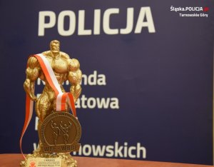 Na zdjęciu widać złoty medal i statuetkę strongmena. W tle znajduje się baner z napisem Policja