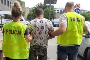 Zdjęcie jest zrobione przed budynkiem policyjnego aresztu, na dziedzińcu Komendy Miejskiej Policji. Zatrzymany, ubrany w koszulkę z krótkim rękawkiem, krótkie spodenki moro ma  ciemną karnację , krótko ostrzyżone włosy, szczupłą budowę ciała. Ma założone na ręce i nogi kajdanki. Po obu jego stronach trzyma go policjantka oraz policjant , ubrani w żółte kamizelki policyjne.&quot;&gt;