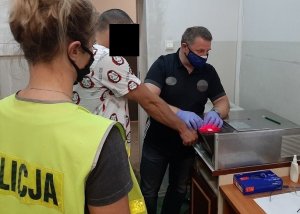 Pomieszczenie w komendzie policji. Technik kryminalistyki daktyloskopuje zatrzymanego przyciskając jego dłoń do urządzenia. Na zdjęciu widać również policjantkę w żółtej kamizelce.&quot;&gt;