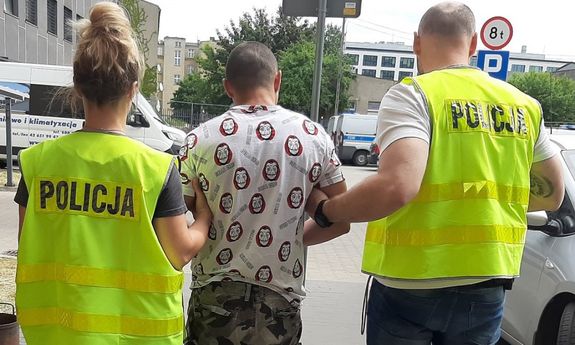 Zdjęcie jest zrobione przed budynkiem policyjnego aresztu, na dziedzińcu Komendy Miejskiej Policji. Zatrzymany, ubrany w koszulkę z krótkim rękawkiem, krótkie spodenki moro ma  ciemną karnację , krótko ostrzyżone włosy, szczupłą budowę ciała. Ma założone na ręce i nogi kajdanki. Po obu jego stronach trzyma go policjantka oraz policjant , ubrani w żółte kamizelki policyjne.&quot;&gt;