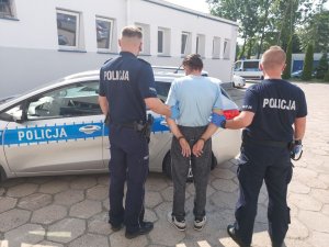 umundurowani policjanci prowadzą zatrzymanego. podejrzany o kradzież ma założone kajdanki na ręce trzymane z tyłu.w tle radiowóz policyjny. zdjęcie zrobione na placu wewnętrznym komendy