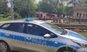 radiowóz policyjny stoi przy stacji kolejowej i przejeździe pociągów, w oddali stoją strażacy przy wozie strażackim