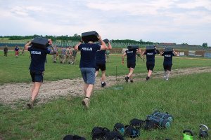 Przenoszenie 10-kilogramowych kanistrów z wodą. Policjanci w czarnych koszulkach niosą na karkach czarne pojemniki.