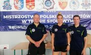 dwóch mężczyzn i kobieta w strojach sportowych na tle baneru Mistrzostwa Polski Policjantów w Tenisie Stołowym