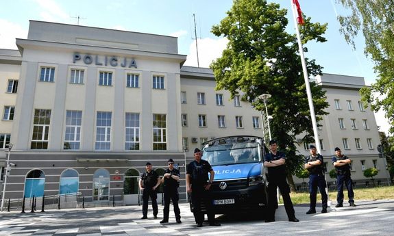 Policjanci stoją przy radiowozie. W tle budynek z napisem Policja