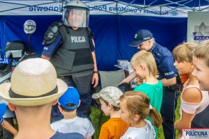 Grupa dzieci stojąca obok Policjant umundurowanego w sprzęt ochrony indywidualnej oraz kask przeciwuderzeniowy z przyłbicą