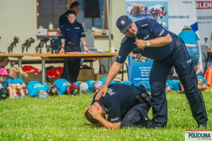 Jeden Policjant klęczy w pozycji obronnej przed psem w pozycji na żółwia, drugi stoi