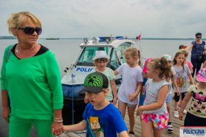 Dzieci wraz z opiekunką stojące na pomoście w tle zacumowana łódź Policyjna