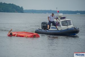 Wywrócona do góry dnem łódka obok mężczyzna w wodzie i stojąca łódź Policyjna z Policjantem na pokładzie
