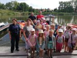 grupa dzieci wraz z opiekunką i policjantką stojących nad wodą za nimi na łodzi policjant z ratownikiem