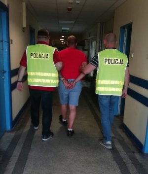 dwaj policjanci , jeden w kamizelce odblaskowej z napisem policja prowadzą zatrzymanego&quot;&gt;