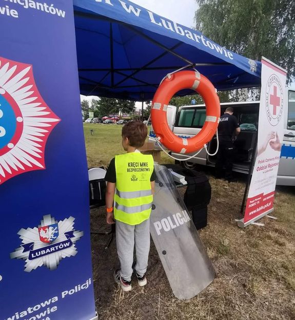 chłopiec odwrócony tyłem stoi pod namiotem Komendy Powiatowej Policji w Lubartowie ma na sobie kamizelkę odblaskową z napisem Kręci mnie bezpieczeństwo obok niego na namiocie zawieszone jest pomarańczowe koło ratunkowe