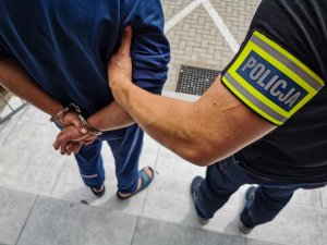 policjant kryminalny w cywilnych ubraniach z założoną żółtą opaską z napisem POLICJA trzyma za ramię mężczyznę podejrzanego o zabójstwo