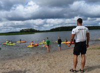 policjant odwrócony tyłem stoi na plaży i czuwa nad bezpieczeństwem grupy dzieci pływajacych na kajakach