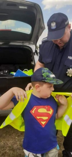 policjant zakłada chłopcu kamizelkę odblaskową z tyłu za nimi otwarty bagażnik radiowozu