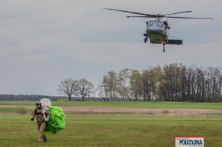 Spadochroniarz opuszcza lądowisko ze zwiniętą czaszą spadochrony na plecach, w tle śmigłowiec podchodzący do lądowania.