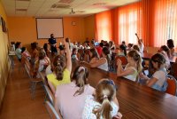 sala wykładowa grupa dzieci siedzących na krzesłach wraz z opiekunem zgłasza się do odpowiedzi na zadawane pytania przez policjantkę