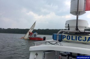 przewrócona żaglówka na jeziorze, która próbują podnieść ze swojej łodzi służby ratunkowe. Z prawej strony, z przodu widać policyjną łódź