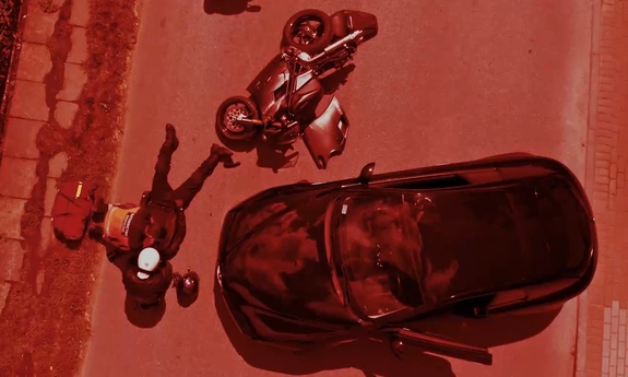 widok z góry na drodze leży motocyklista obok niego siedzi drugi obok dwa motory  jeden z nich przewrócony z boku stoi samochód