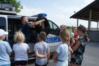 policjant obok radiowozu zakłada dziecku kamizelkę policyjną obok stoją inne dzieci