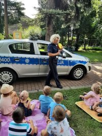 radiowóz obok policjantka z grupą siedzących na trawie przedszkolaków