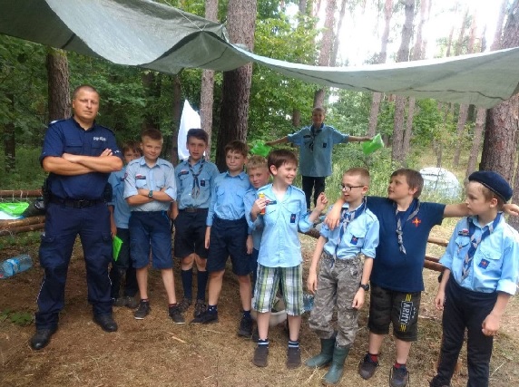 Policjant w mundurze stojący w lesie wraz z dziećmi ubranymi w bluzy harcerskie