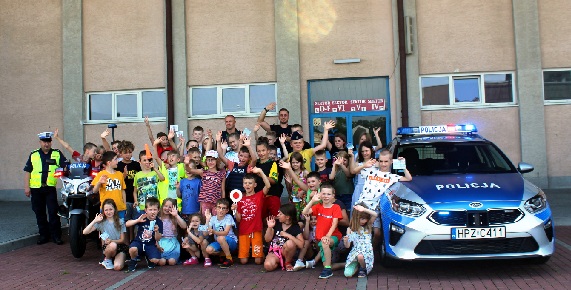 Policjanci w mundurach stoją wraz z młodzieżą na zdjęciu grupowym obok radiowozu.