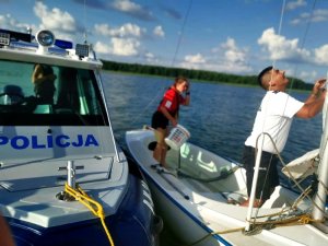Łódź policyjna przy jachcie na jeziorze, policjant i ratownik WOPR na odwróconej łodzi