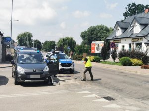 Ulica w mieście. Dwaj ubrani po cywilnemu policjanci stoją na ulicy przy otwartym samochodzie. Z tyłu widoczny oznakowany policyjny radiowóz