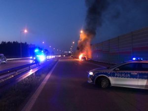 zdjęcie miejsca zdarzenia. Na pierwszym planie oznakowany radiowóz z włączonymi sygnałami świetlnymi w oddali pojazd straży pożarnej również z włączonymi światłami uprzywilejowania. Po prawej stronie drogi pojazd objęty ogniem, widoczne płomienie i kłęby dymu nad pojazdem