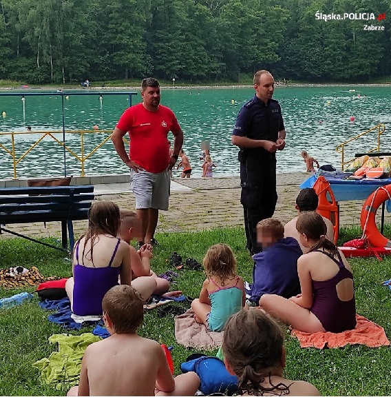 Policjant oraz Ratownik WOPR stojący przed grupą dzieci, które siedzą na trawie