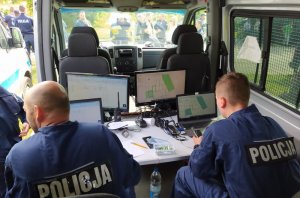 Centrum dowodzenia w jednym z radiowozów. Przed monitorami siedzi dwóch policjantów. Na monitorach widok map z zaznaczonymi sektorami do poszukiwań