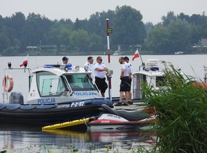 Umundurowani policjanci stojący na pomoście, gdzie obok zacumowane są pontony motorowe Policji oraz skuter WOPR z zaczepioną z tyłu żółtą deską ratowniczą.