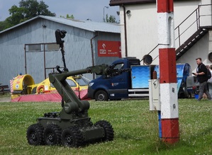 Robot rozpoznania saperskiego, specjalnym chwytakiem zdejmuje ze skrzynki energetycznej, przymocowanej do biało czerwonego słupa, imitację granatu - ładunku wybuchowego.