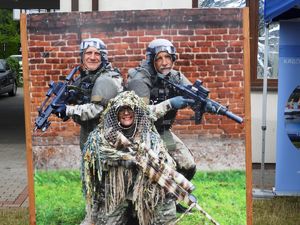 Drewniana płyta z nadrukowaną fotografią trzech osób w umundurowaniu taktycznym i maskującym Policji z bronią z wyciętymi zamiast twarzy otworami. W otworach widać twarze dwóch mężczyzn i kobiety.
