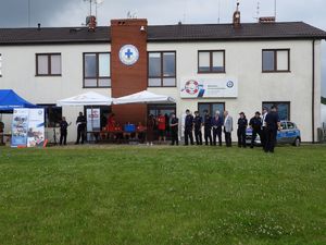 Trawiasty plac z jednopiętrowym budynkiem z jasną elewacją i brązowym dachem, na którym w środkowej części umieszczono oficjalne logo WOPR. Na zdjęciu widać kilkanaścioro ludzi. Większość z nich ubrana jest w granatowe policyjne mundury. Bezpośrednio za nimi stoi oznakowany radiowóz policyjny.