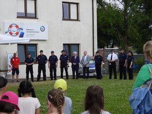 Trawiasty plac z jednopiętrowym budynkiem z jasną elewacją i banerem PZU z napisem „Wpływamy na bezpieczeństwo”. Na zdjęciu widać kilkanaścioro ludzi. Większość z nich ubrana jest w granatowe policyjne mundury. Bezpośrednio za nimi stoi oznakowany radiowóz policyjny. Na pierwszym planie widać głowy kilkorga dzieci.