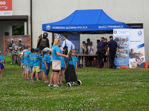 Dzieci ubrane w niebieskie koszulki z napisem Wieliszew PS1 W tle granatowy namiot z napisem Komenda Główna Policji Biuro Prewencji obok którego stoją umundurowani Policjanci. Po bokach namiotu stoi manekin i banery Policji i PZU.