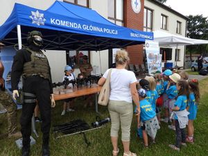 Dzieci ubrane w niebieskie koszulki z napisem Wieliszew PS1 W tle granatowy namiot z napisem Komenda Główna Policji Biuro Prewencji pod którym na stołach leżą różne jednostki broni palnej długiej. W tle stoi radiowóz policyjny i umundurowani Policjanci. Po bokach namiotu stoi manekin i baner Policji.